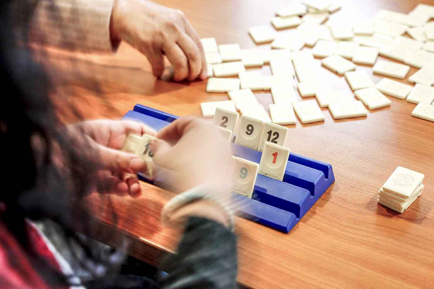 Zwei Personen spielen das Gesellschaftsspiel Rummikub an einem Holztisch. Im Vordergrund ist eine Hand beim Platzieren eines Spielsteins auf einem blauen Spielbrett zu sehen, während im Hintergrund zahlreiche weitere Spielsteine auf dem Tisch verteilt sind. Die Spielsteine zeigen Zahlen in verschiedenen Farben.