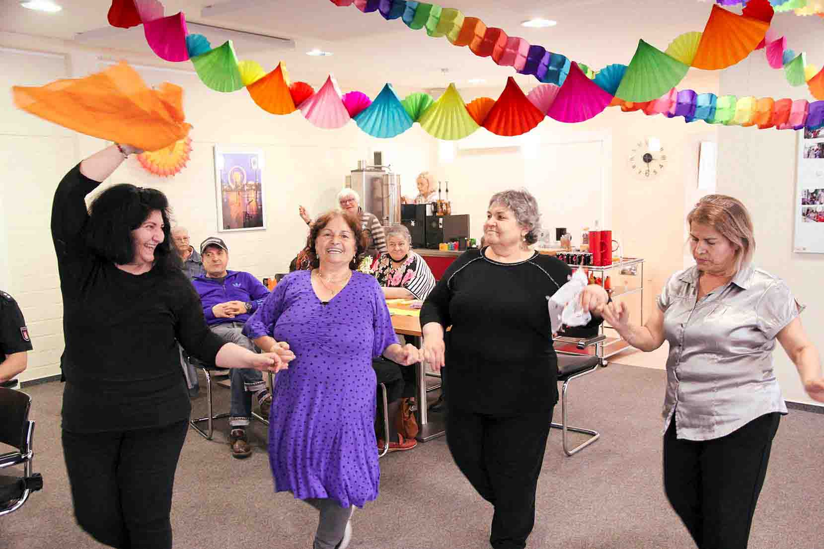 Eine Gruppe älterer Frauen tanzt fröhlich in einem festlich geschmückten Raum. Die Frauen halten sich an den Händen und lächeln, während sie bunte Tücher in der Luft schwingen. Im Hintergrund sitzen weitere Seniorinnen und Senioren an einem Tisch und schauen zu. Die Decke ist mit bunten Papiergirlanden dekoriert, was dem Raum eine festliche und fröhliche Atmosphäre verleiht.