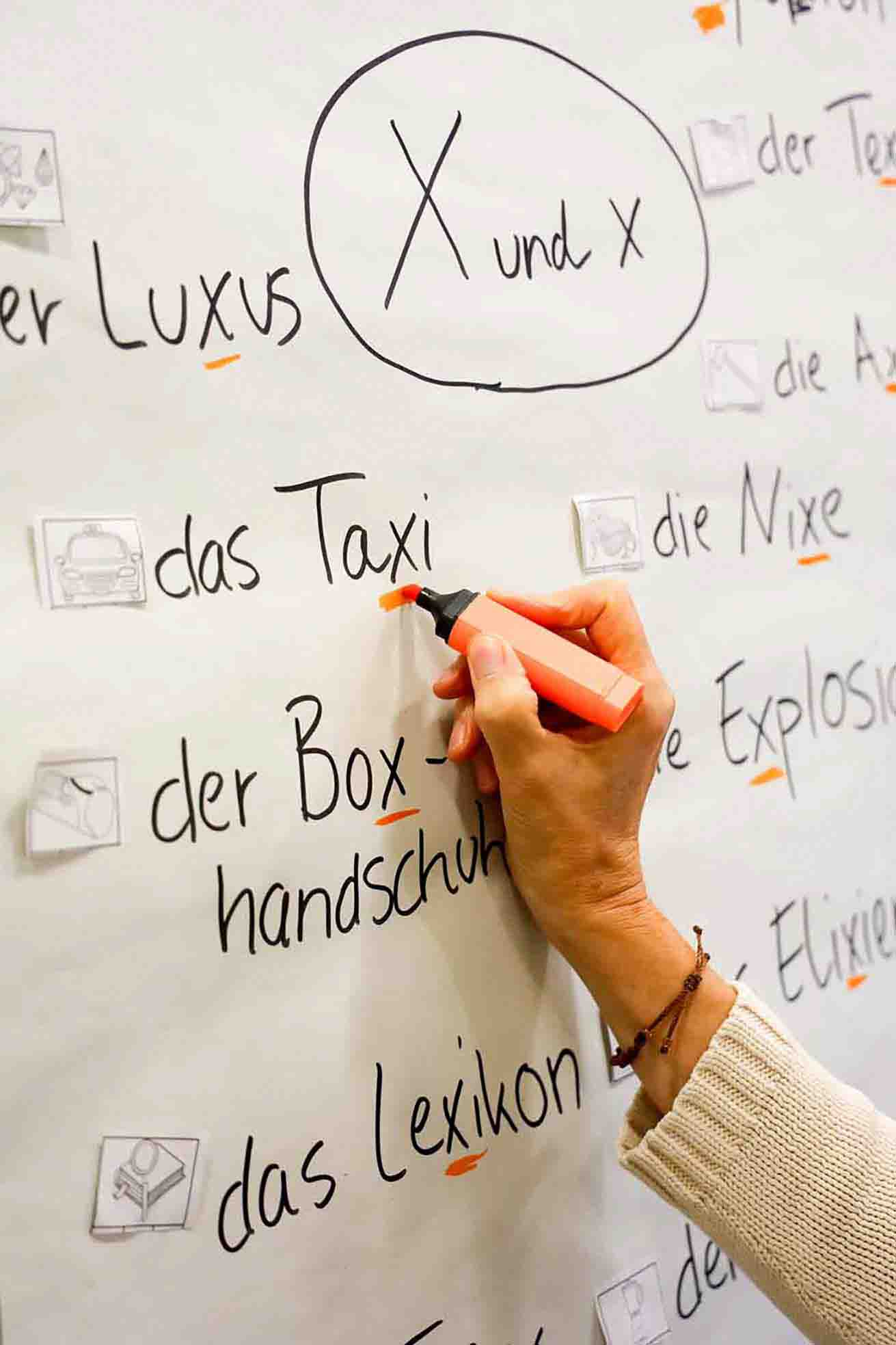 Eine Person markiert mit einem orangefarbenen Textmarker das Wort „Taxi“ auf einem großen Lernplakat. Auf dem Plakat stehen verschiedene deutsche Wörter, die den Buchstaben X enthalten, jeweils mit Artikel und kleinen Symbolbildern versehen. Im oberen Bereich ist „X und x“ in einem Kreis hervorgehoben.