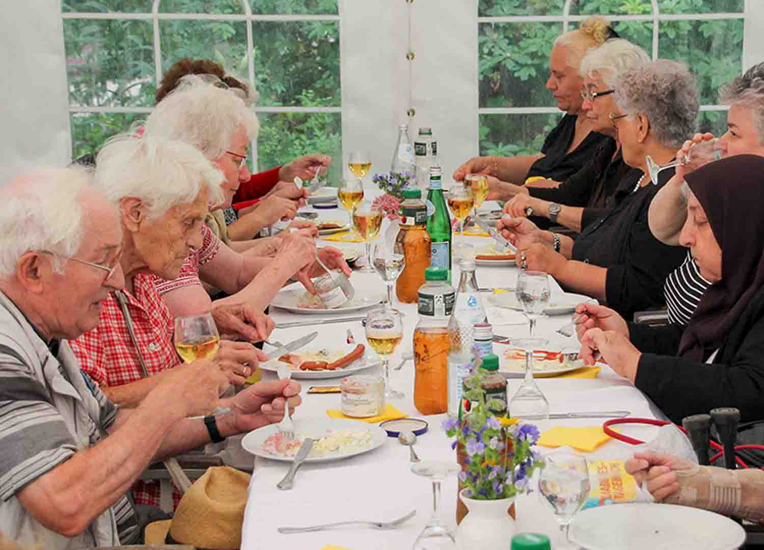 Seniorengruppe sitzt gemeinsam an einer langen, festlich gedeckten Tafel unter einem weißen Zelt und isst zusammen.