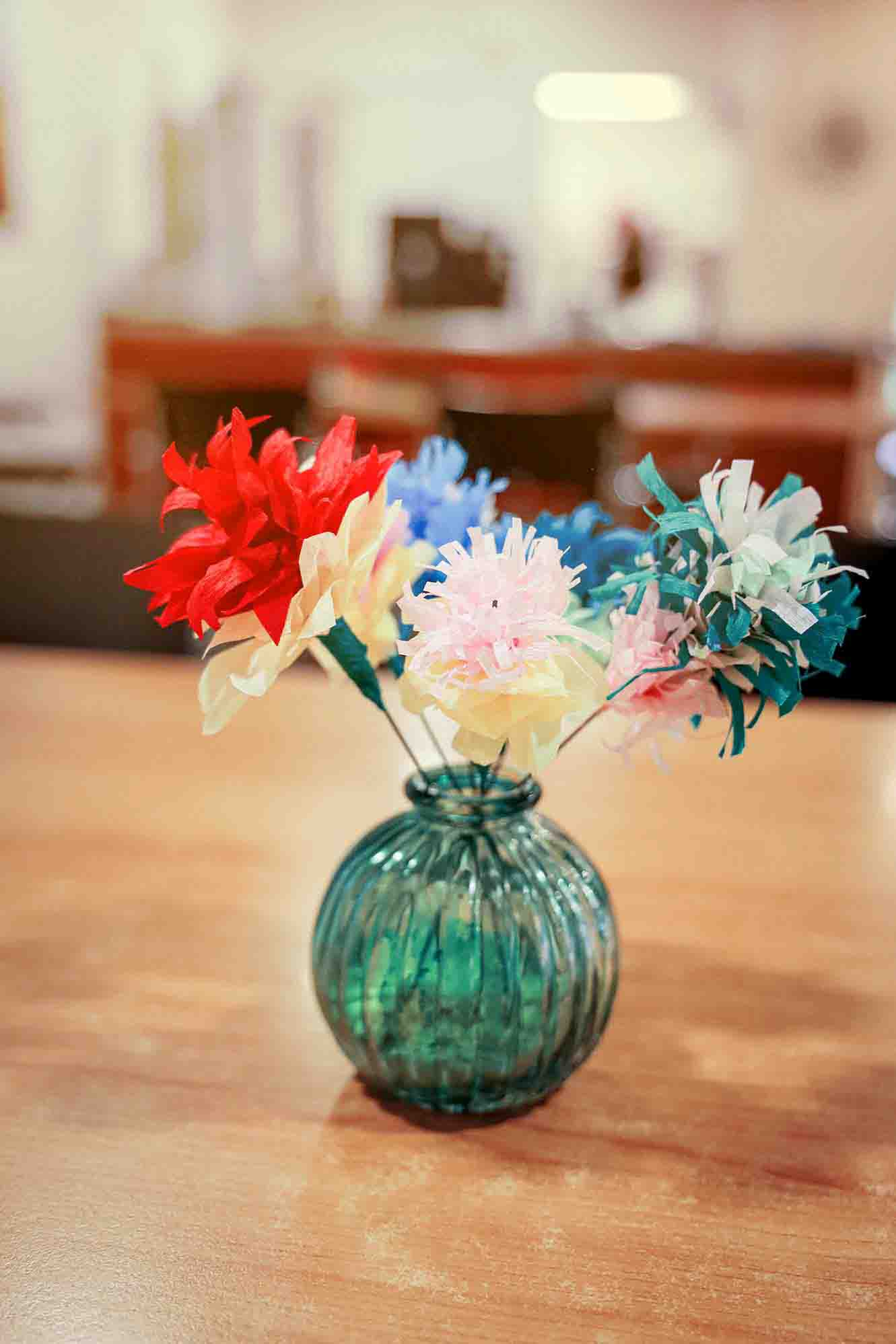 Bunte Papierblumen in einer geriffelten, grünen Glasvase stehen auf einem Holztisch in einem hellen Raum.