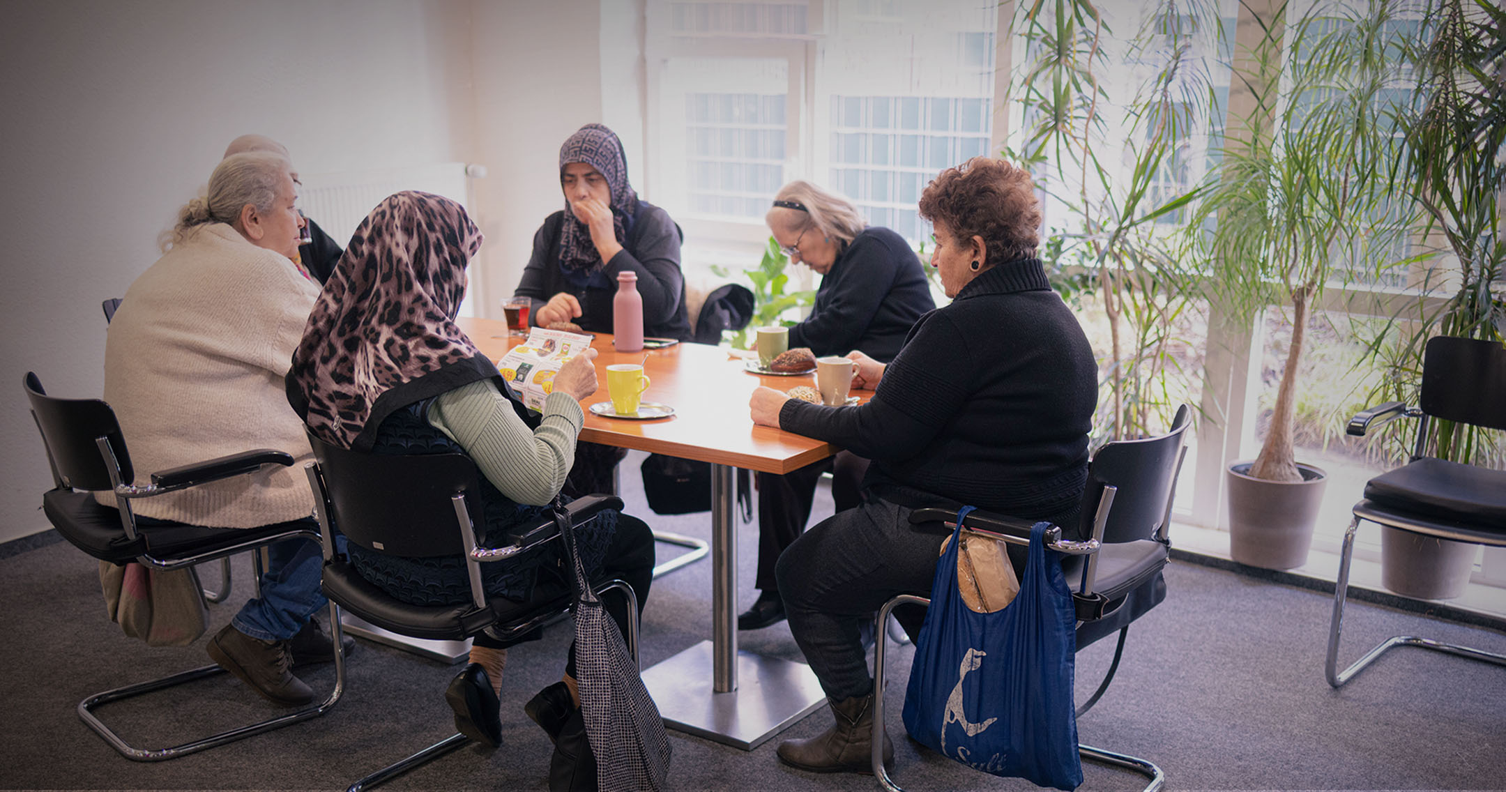 Sechs ältere Menschen, darunter Frauen mit und ohne Kopftuch, sitzen an einem Tisch in einem hellen Gemeinschaftsraum mit großen Fenstern und Topfpflanzen. Sie unterhalten sich, trinken Tee oder Kaffee und lesen.