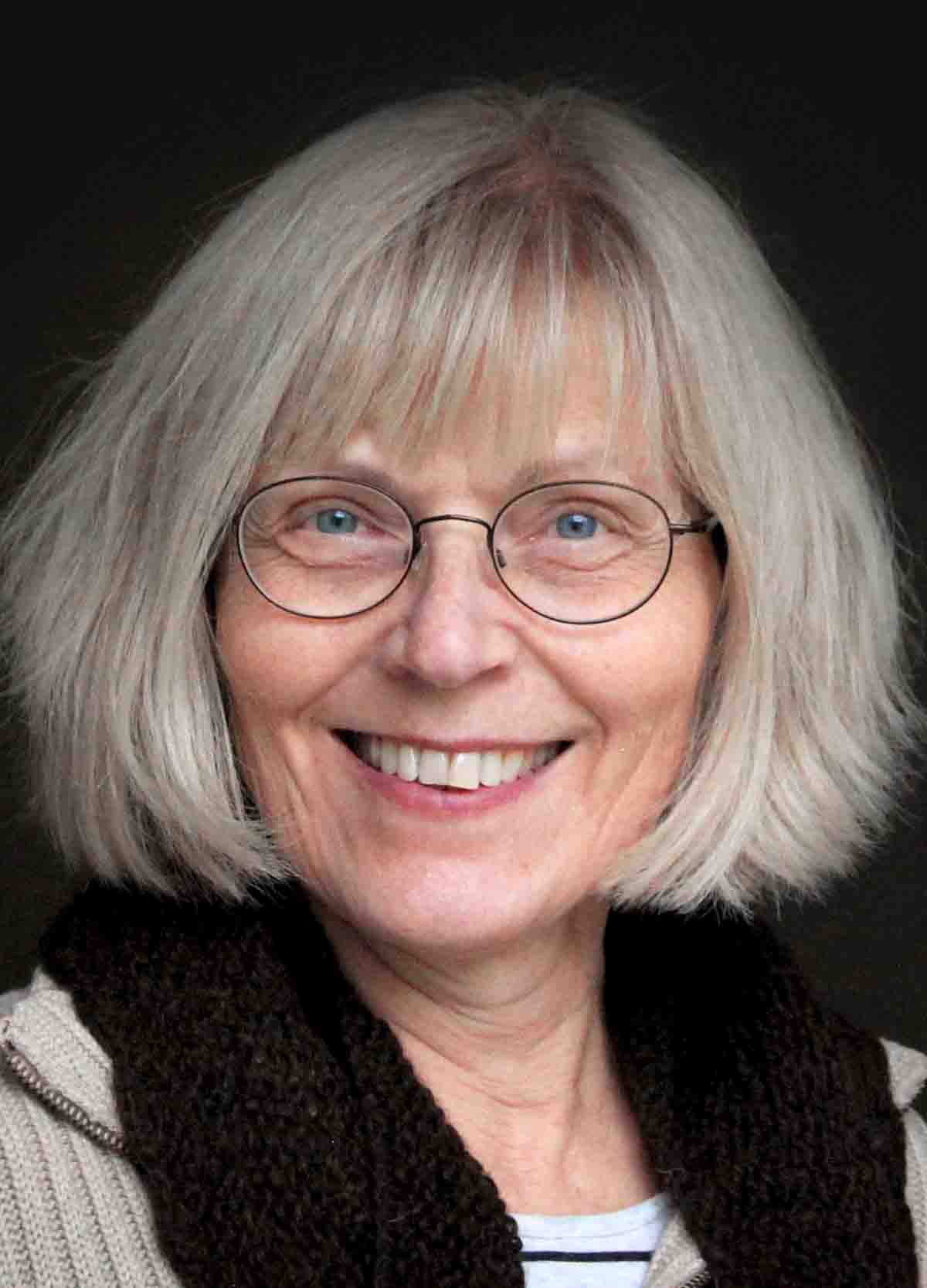 Porträt einer Frau mit grauem, schulterlangem Haar und Brille, die freundlich in die Kamera lächelt. Sie trägt einen gestrickten Pullover mit einem dunklen Schal.
