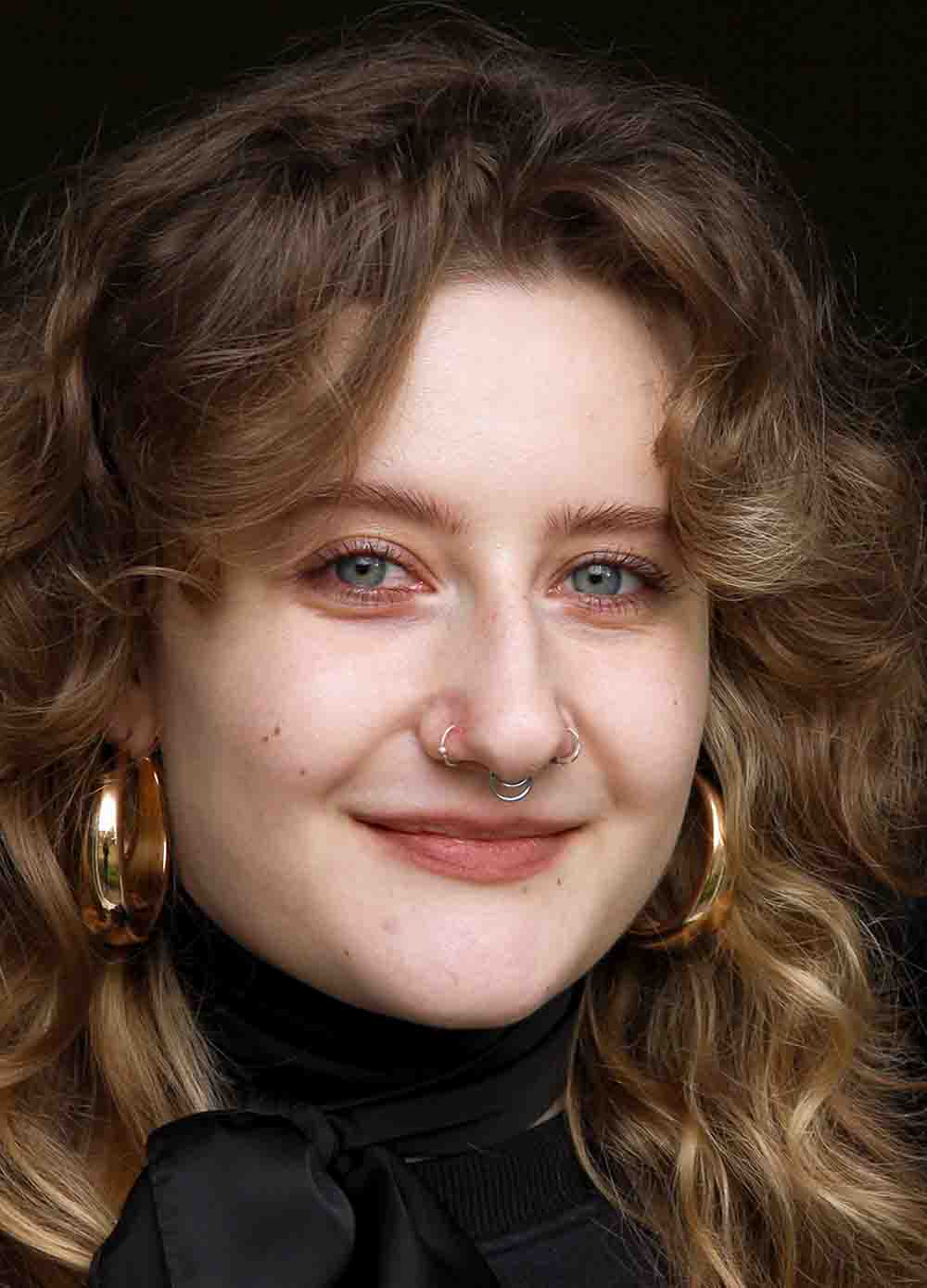 Porträt einer jungen Frau mit lockigem Haar, Nasenpiercings und goldenen Ohrringen, die freundlich lächelt